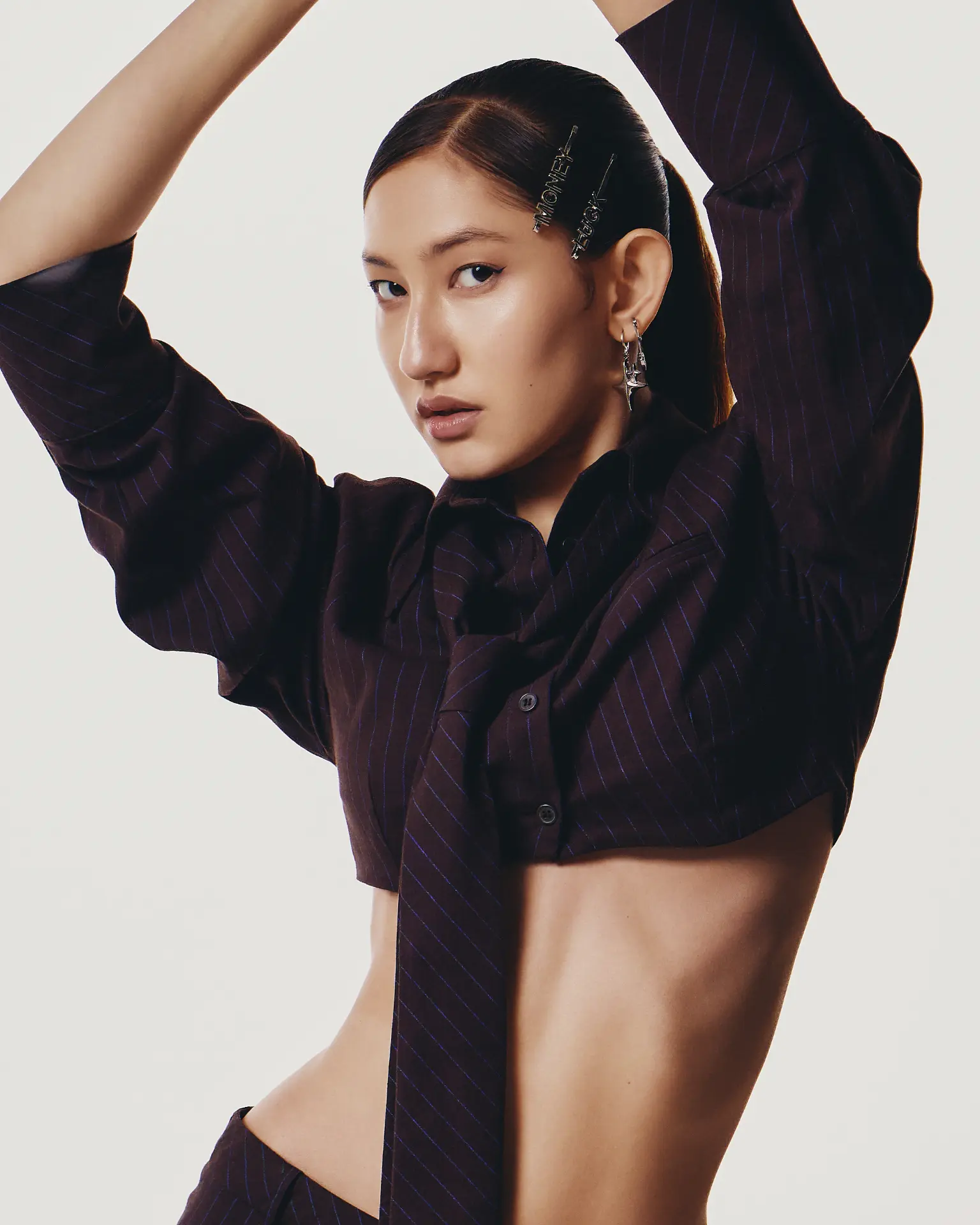 Portrait of a woman in a pinstriped cropped shirt with her arms raised, looking at the camera