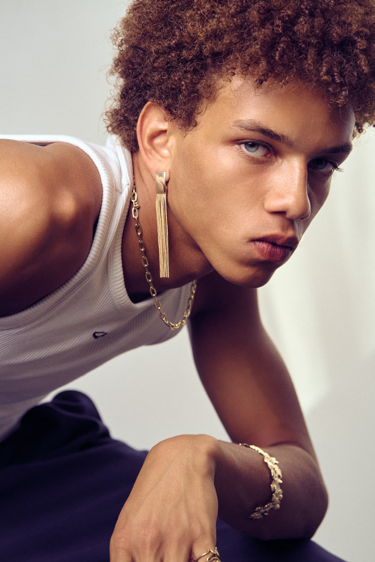 Close-up portrait of a curly-haired man wearing a gold earring and chain, leaning forward