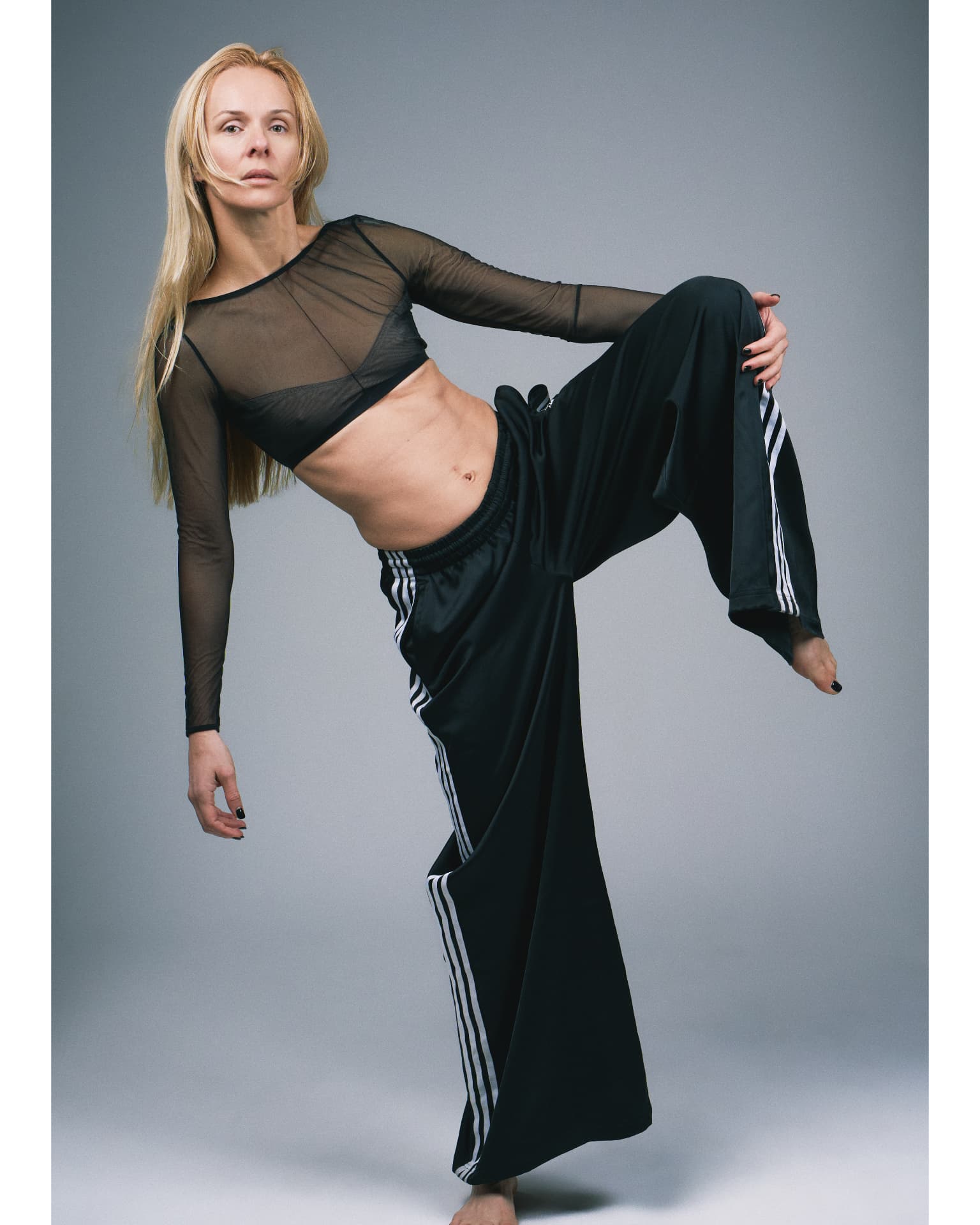 Full-length portrait of a blonde woman in a sheer black top and striped trousers holding her leg raised