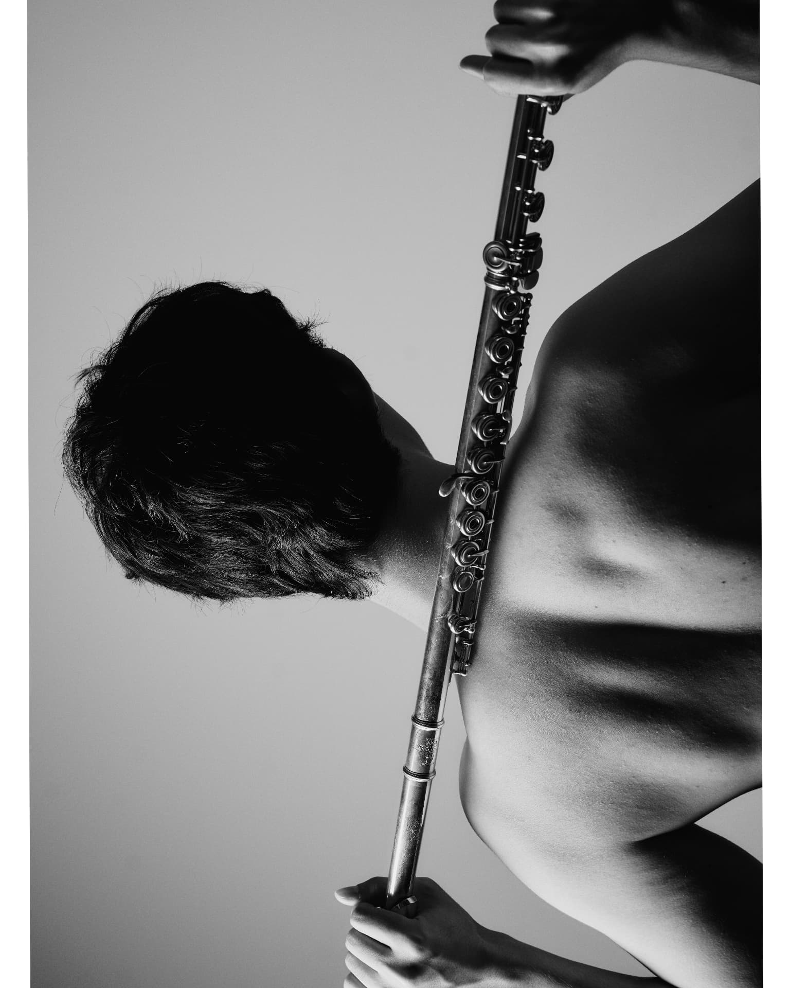 Black-and-white rotated portrait of a shirtless man holding a flute vertically