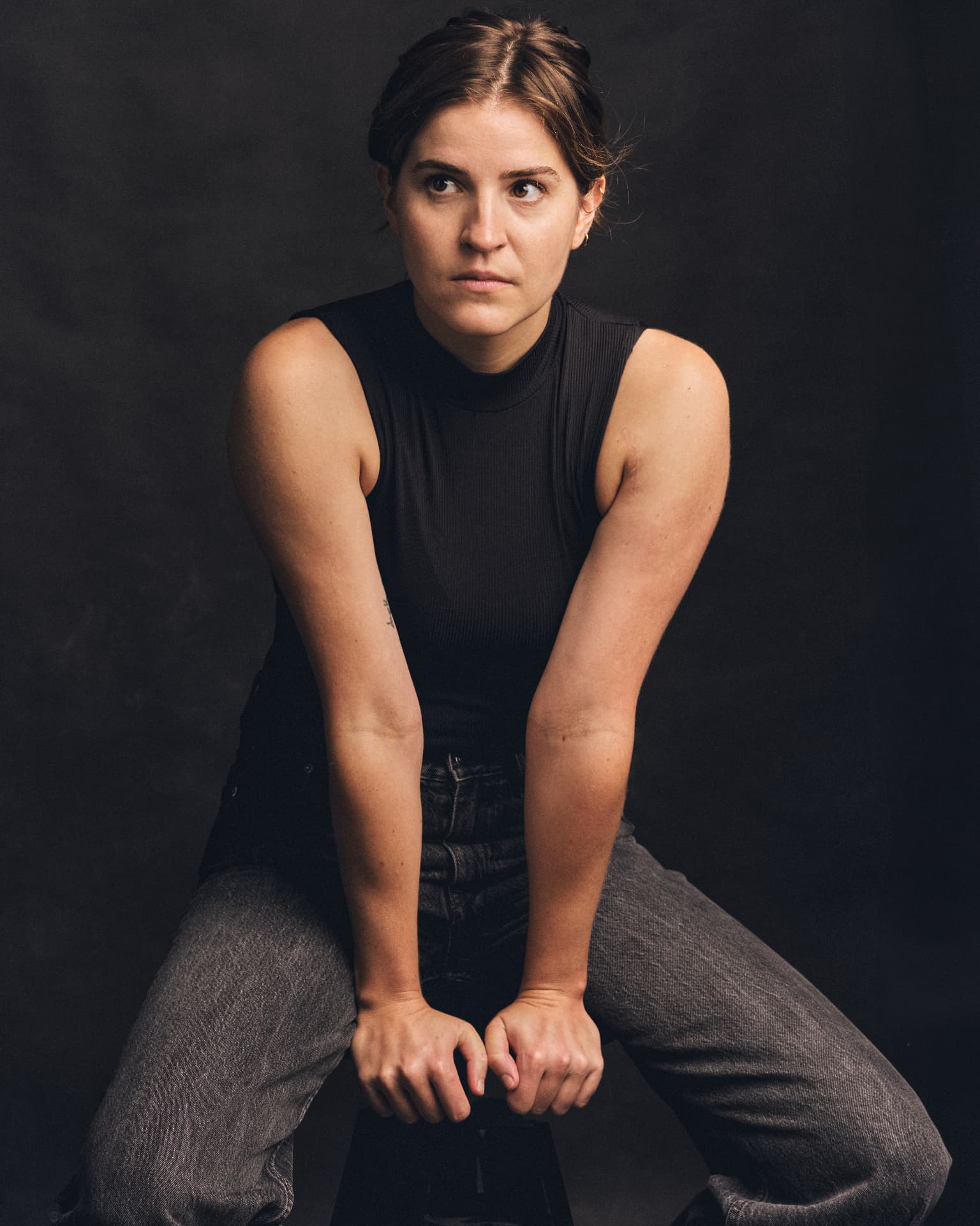 Portrait of a woman seated on a stool in a black top, against a dark background