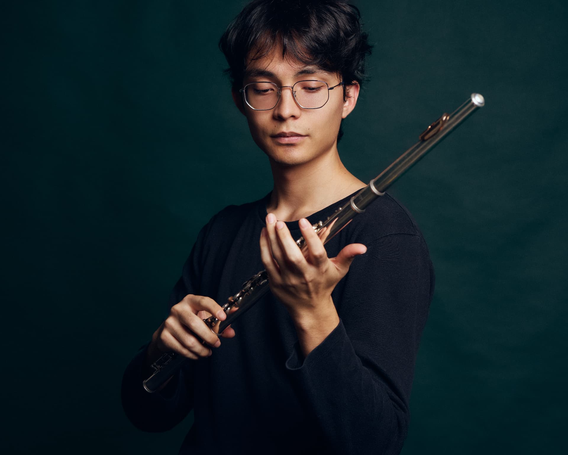 Portrait of a young man in glasses holding a flute against a dark green background, eyes closed