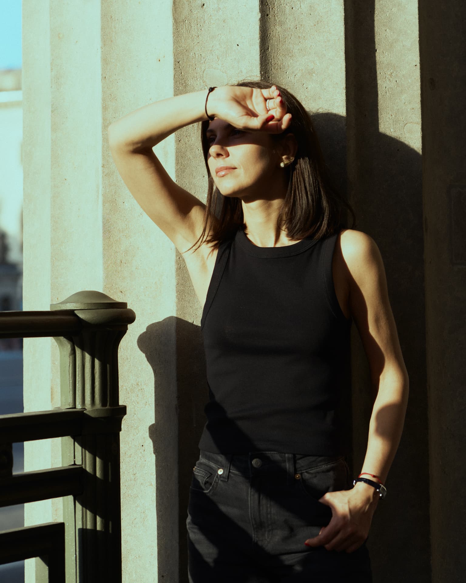 Sunlit portrait of a woman in a black tank top shading her eyes against a concrete wall
