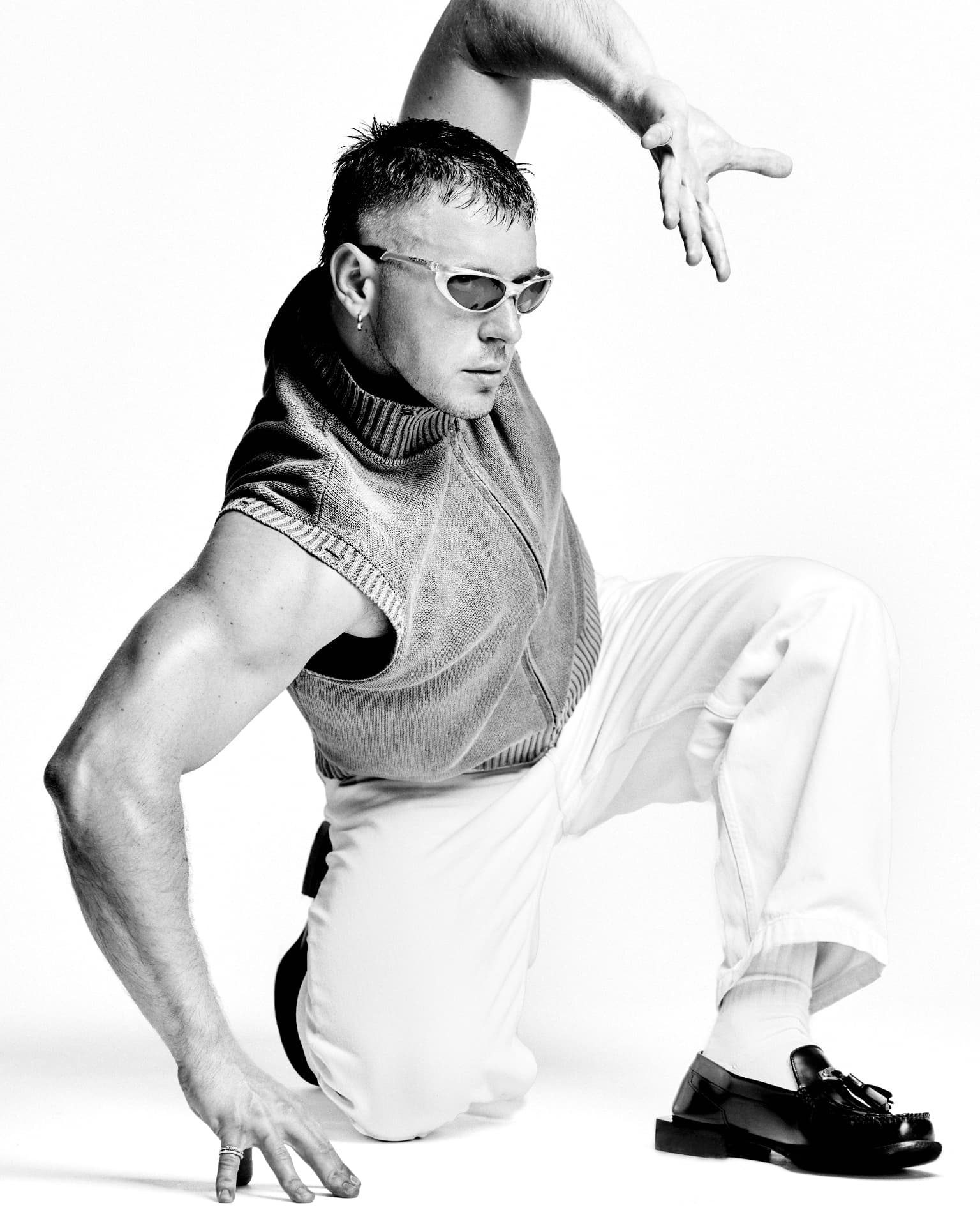 Black-and-white full-length portrait of a man in sunglasses crouching low in a knit vest and white trousers