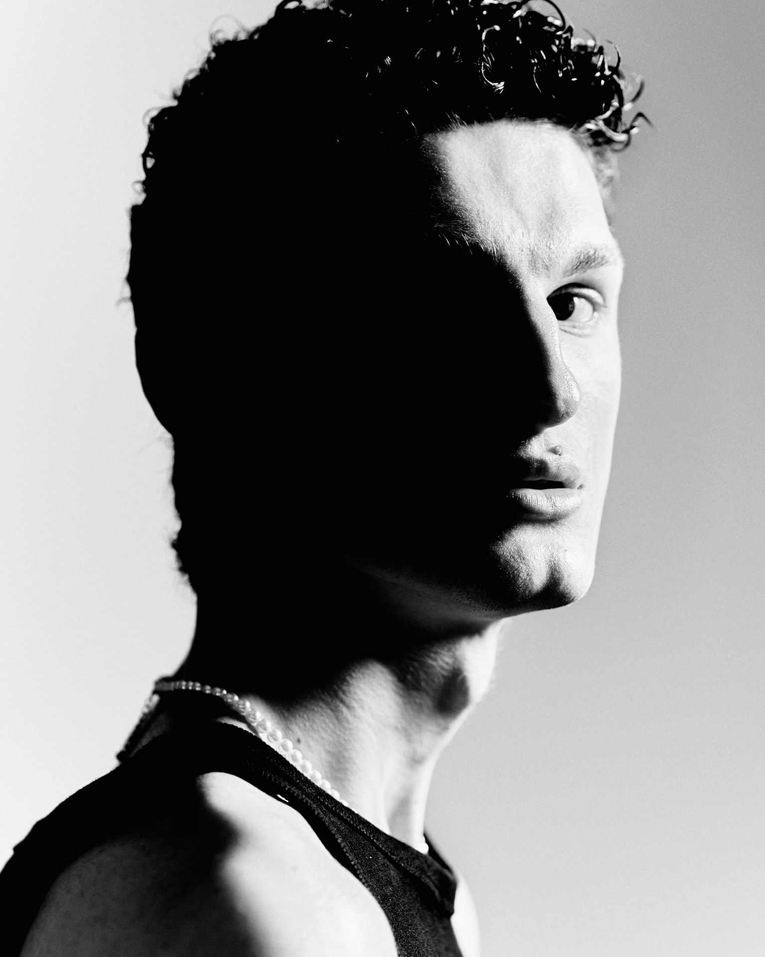 Black-and-white close-up portrait of a curly-haired man in a tank top and pearl necklace, face half in shadow