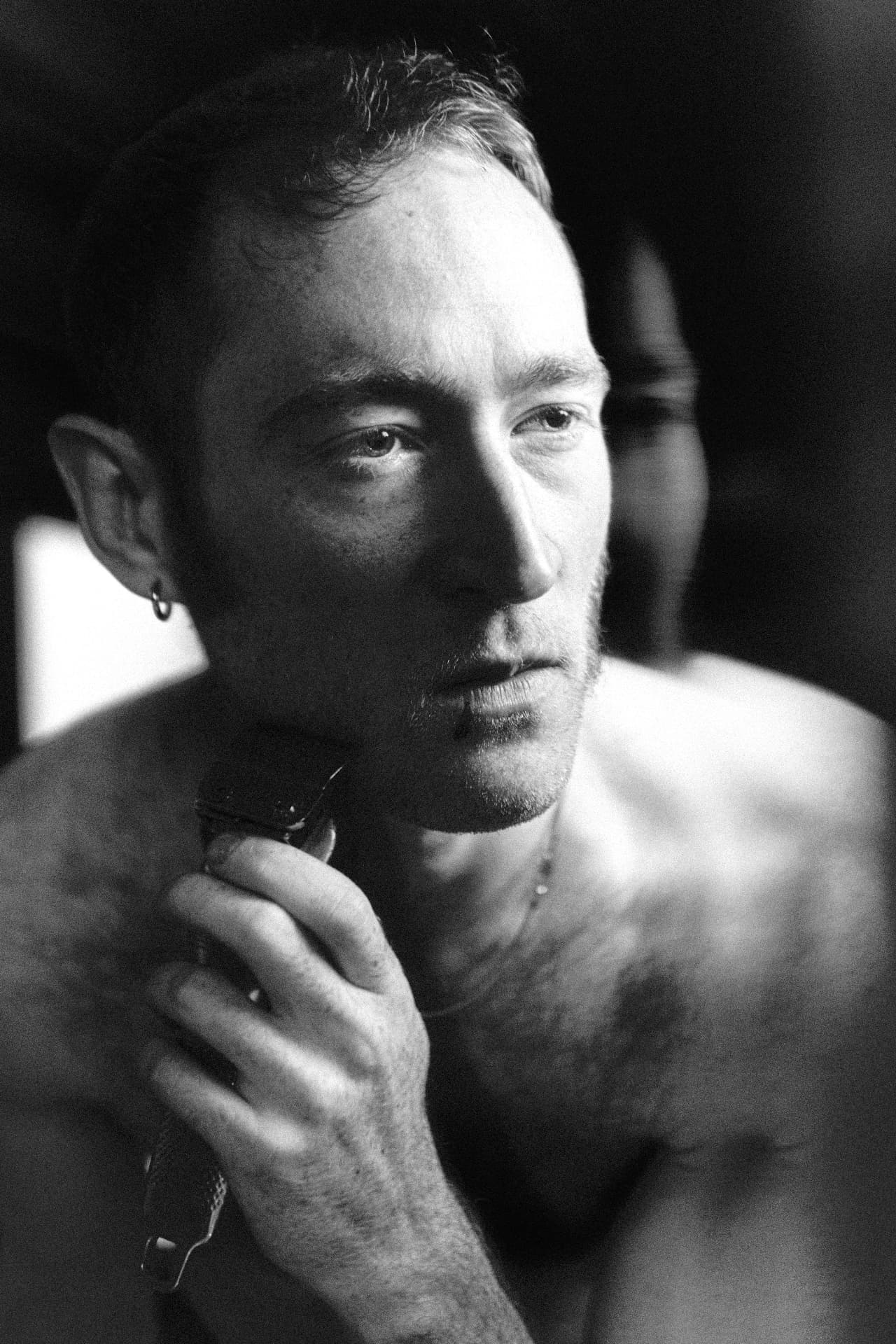 Black-and-white close-up of a shirtless man holding a hair clipper near his face