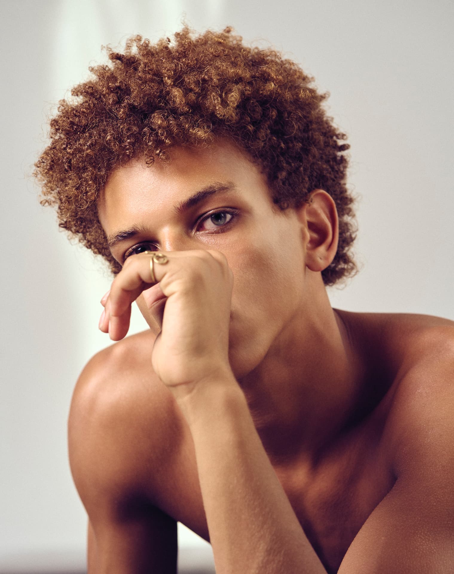 Close-up portrait of a shirtless curly-haired man resting his hand against his mouth