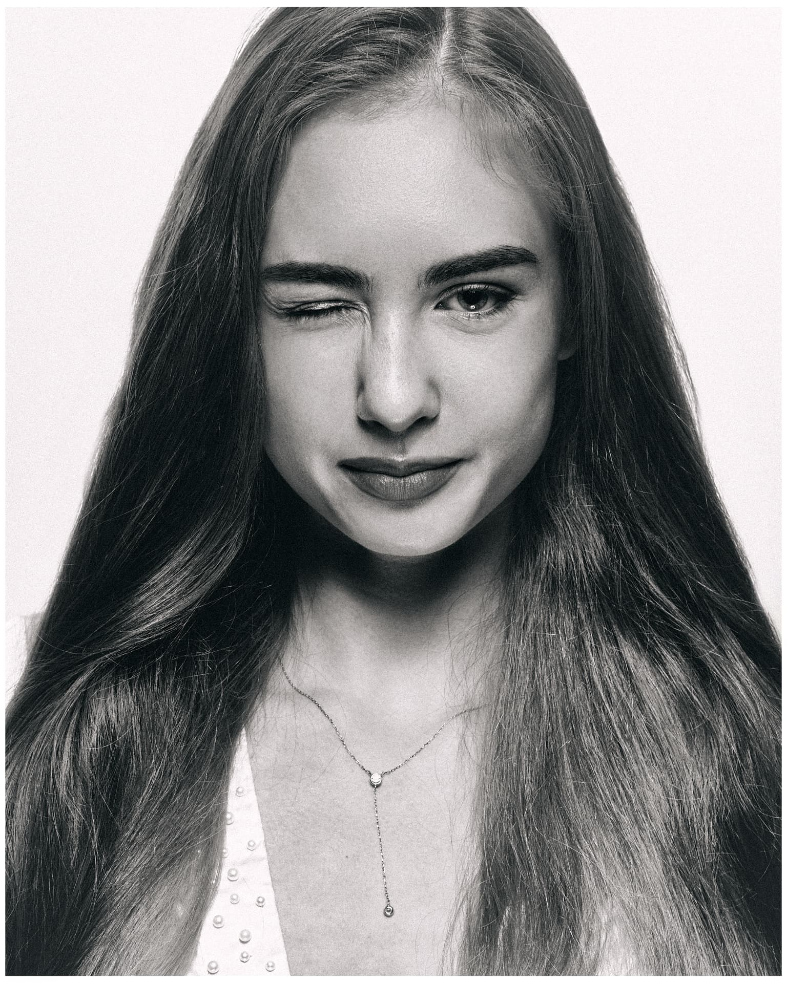 Black-and-white close-up portrait of a young woman winking, with long hair and a delicate necklace