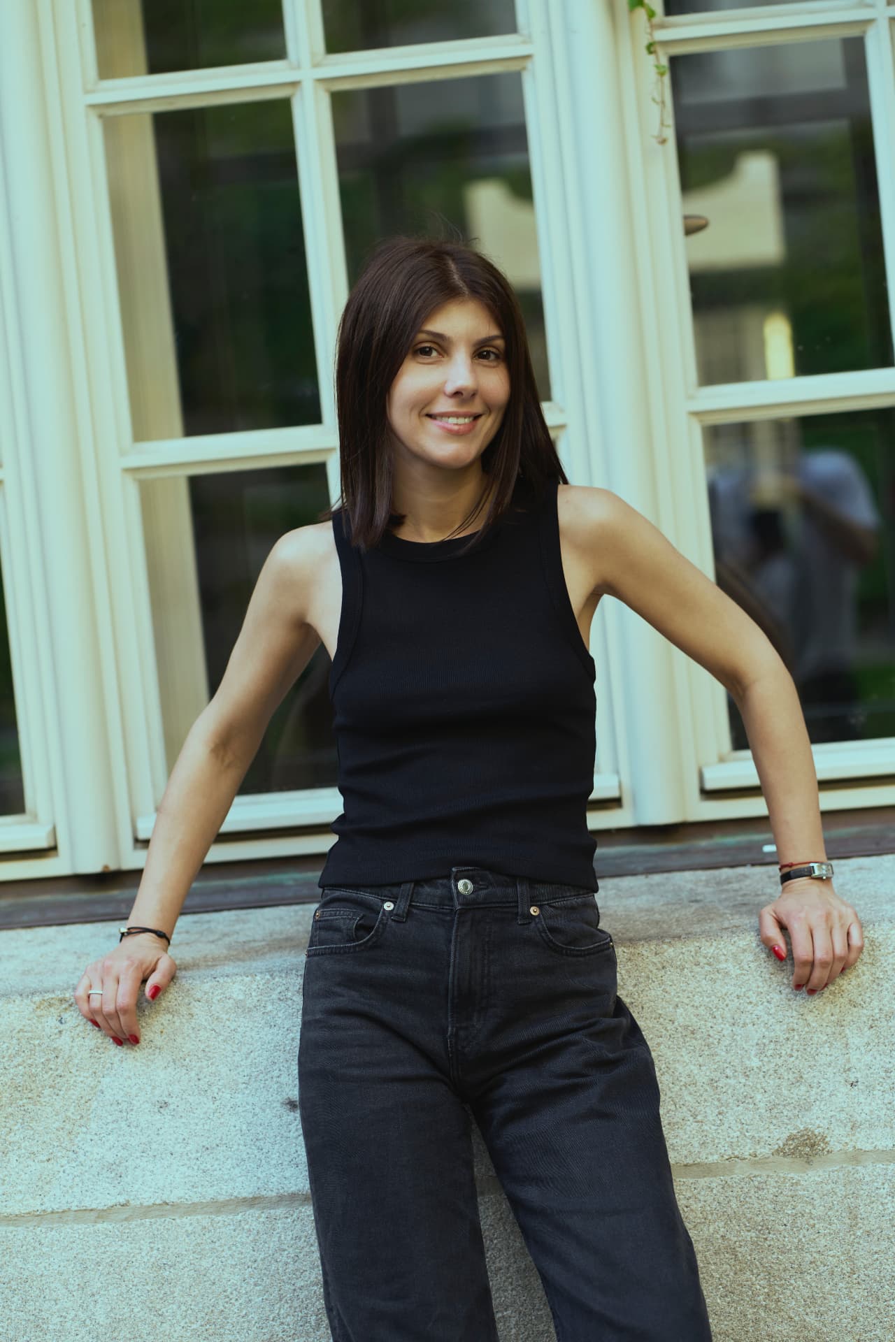 Portrait of a woman in a black tank top and jeans leaning against a window frame outdoors