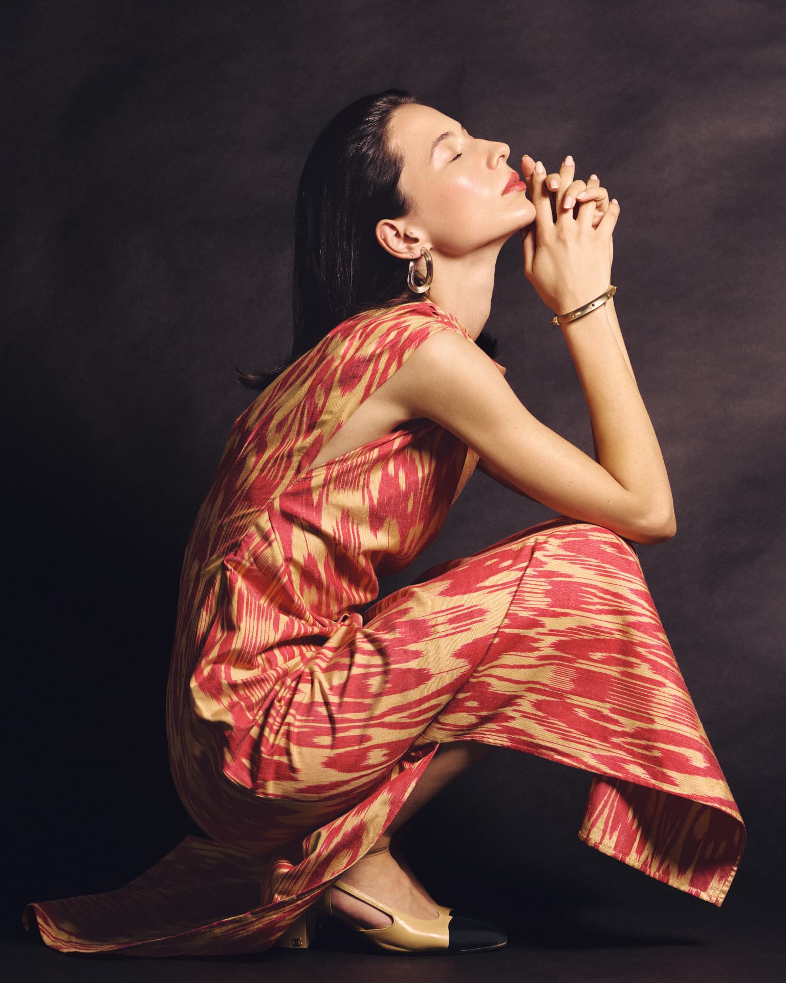 Full-length portrait of a woman crouching in a red and gold patterned dress, eyes closed