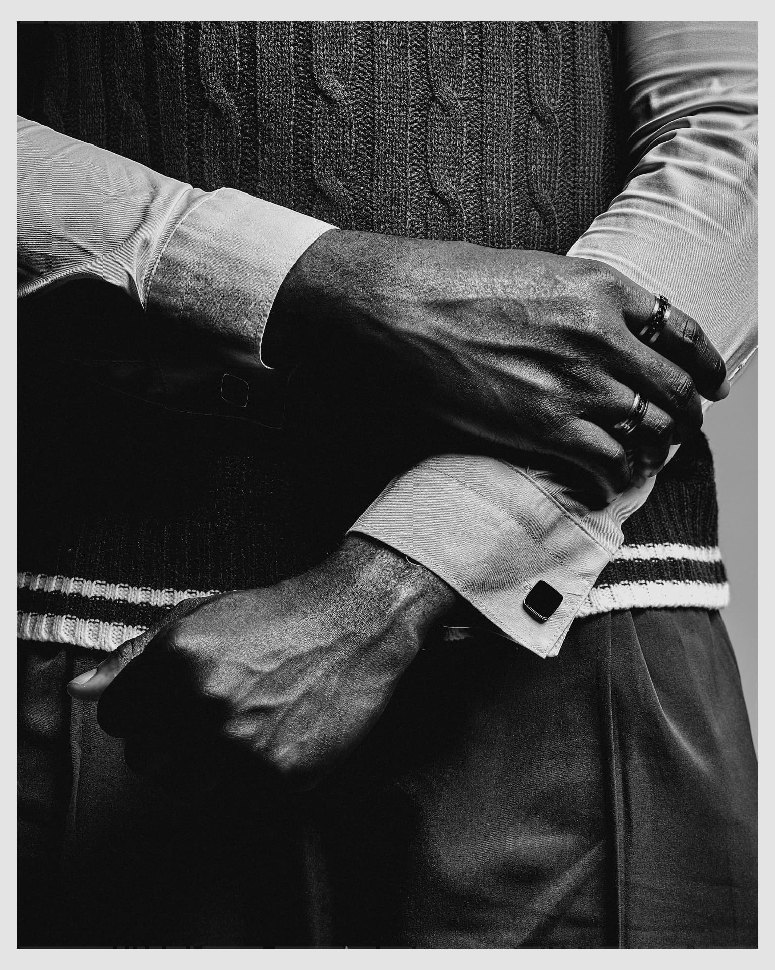 Black-and-white detail of a man's hands fastening his cufflinks over a cable-knit vest