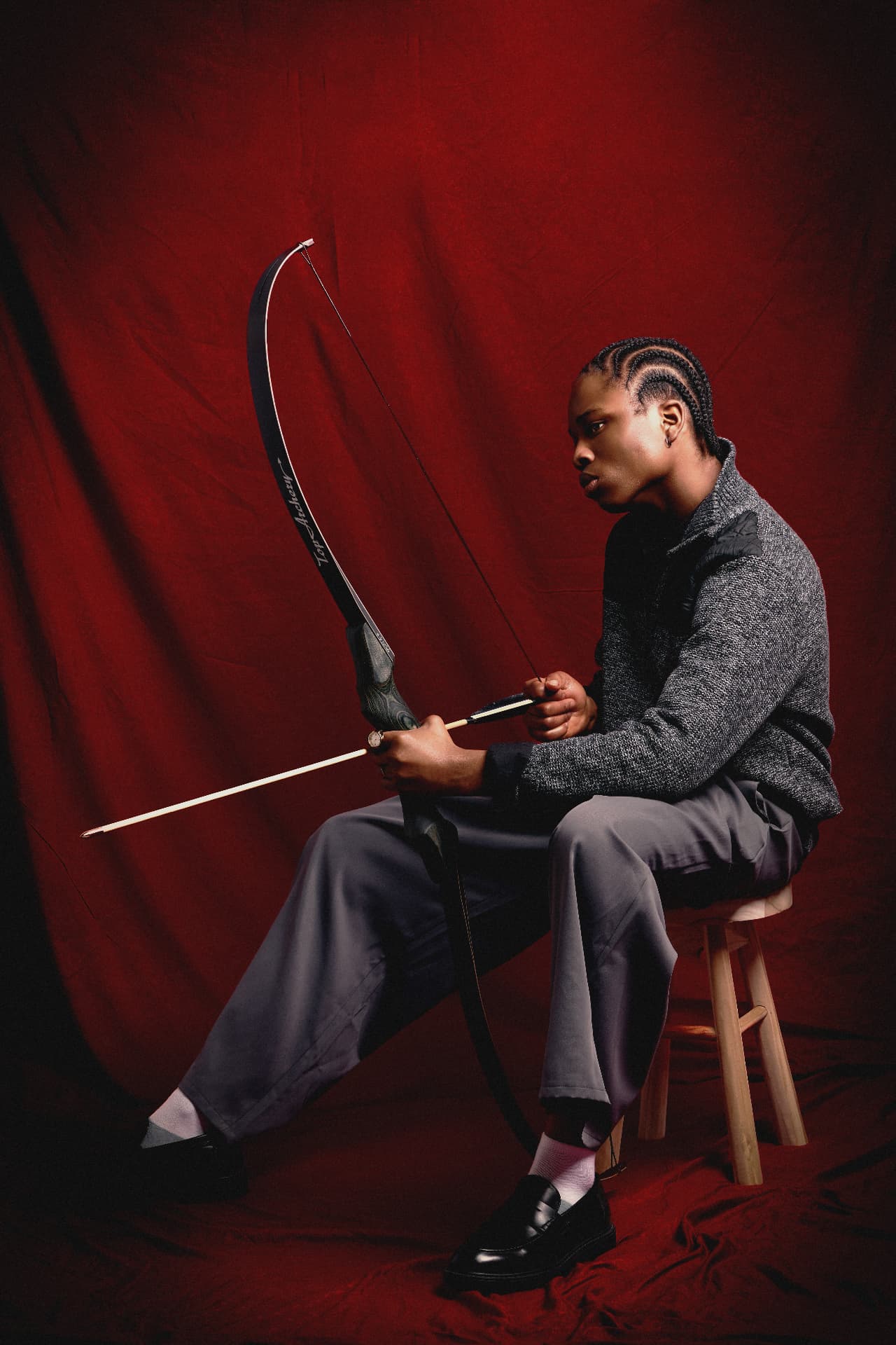 Portrait of a person with braided hair holding a bow and arrow, seated against a red backdrop