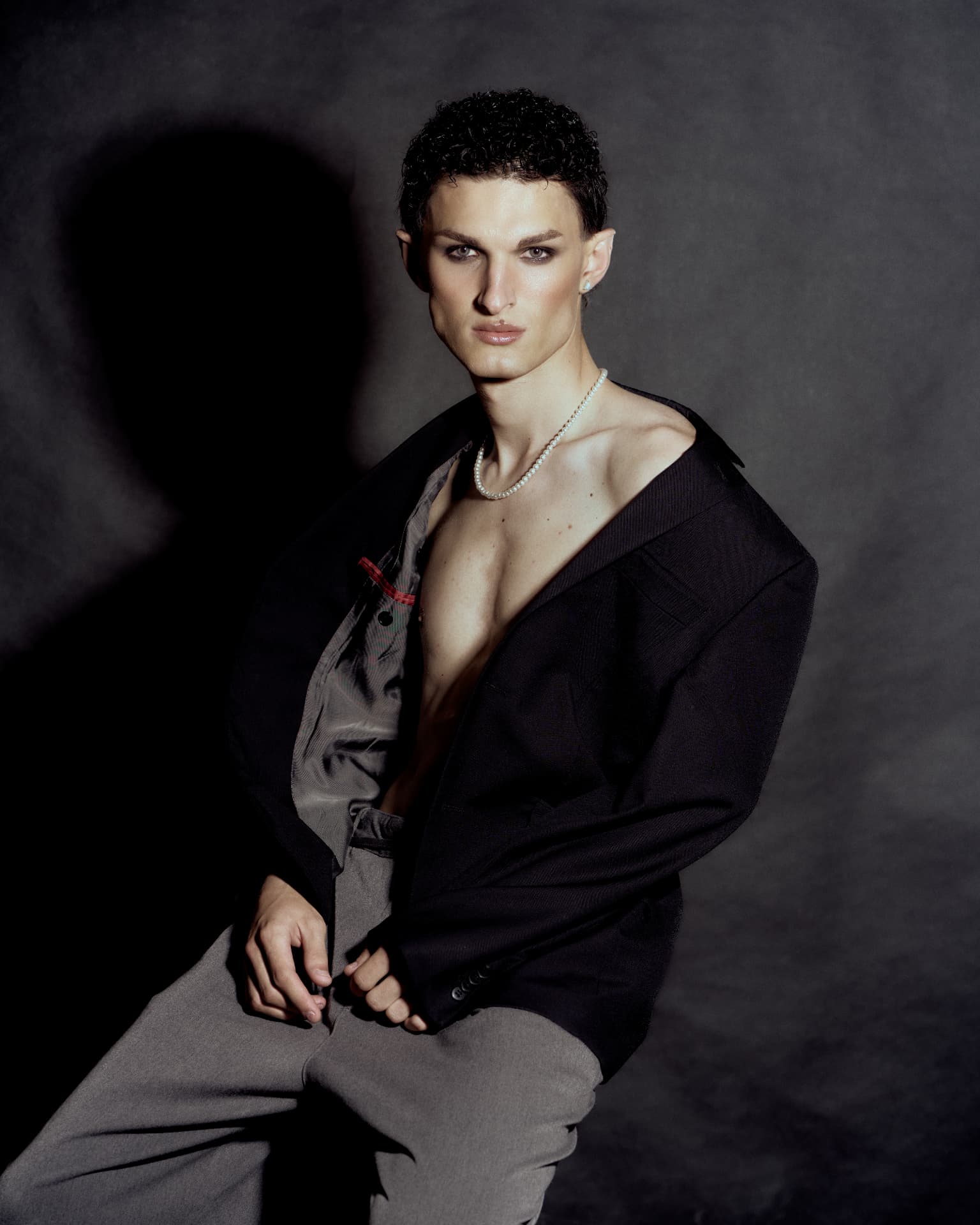 Portrait of a curly-haired man in an open black jacket and pearl necklace, seated against a dark backdrop