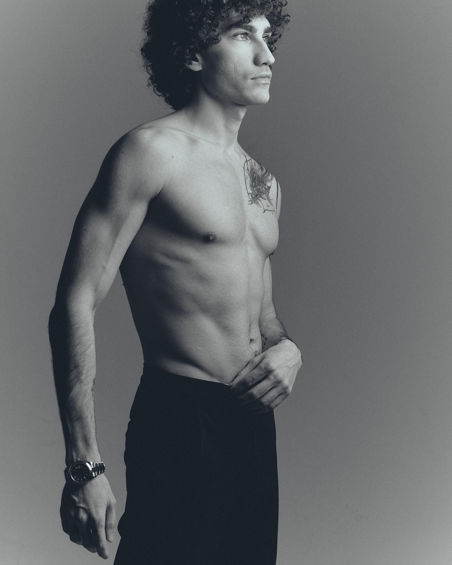 Black-and-white profile portrait of a shirtless man with curly hair and black trousers