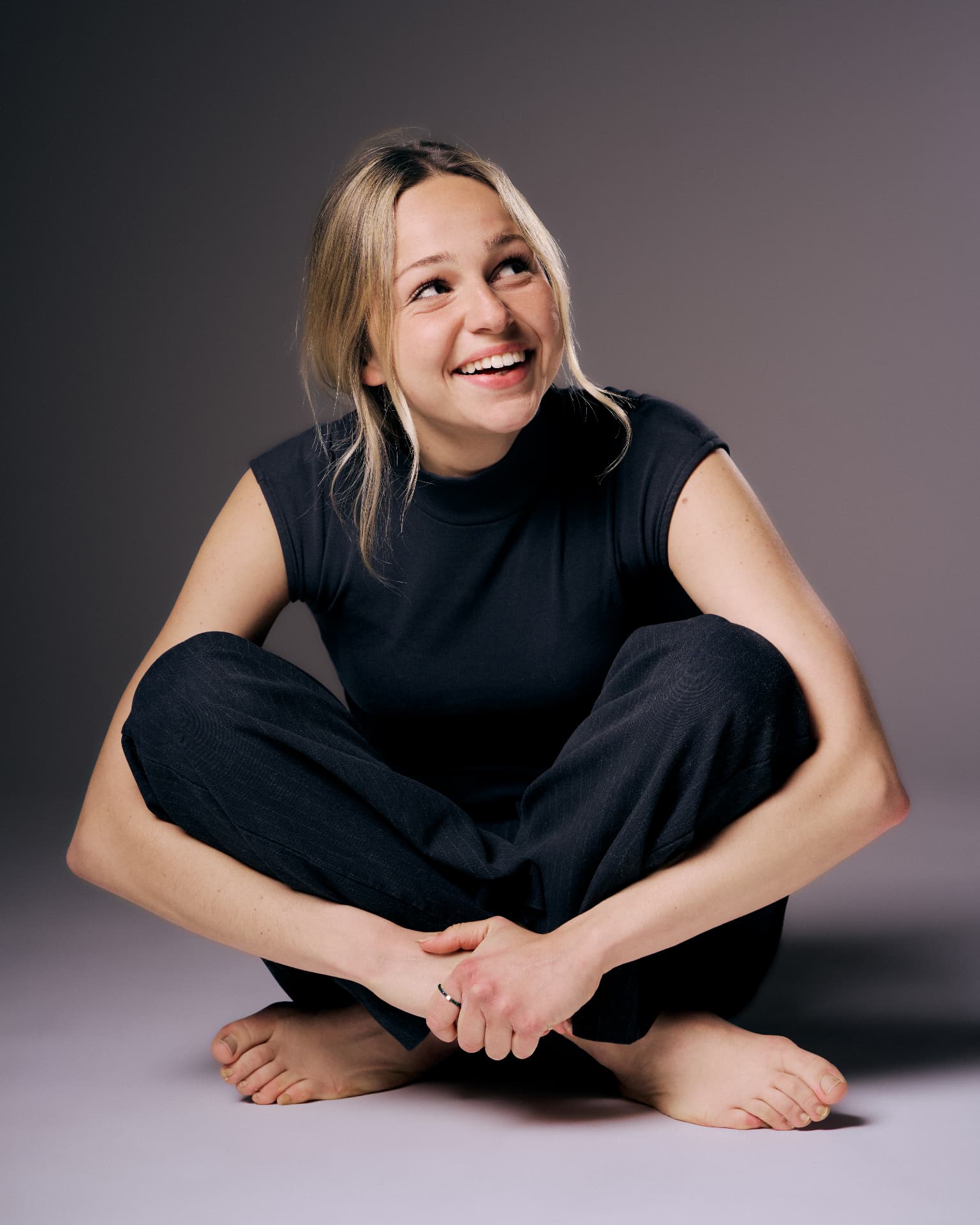Portrait of a smiling blonde woman seated cross-legged in black, looking up