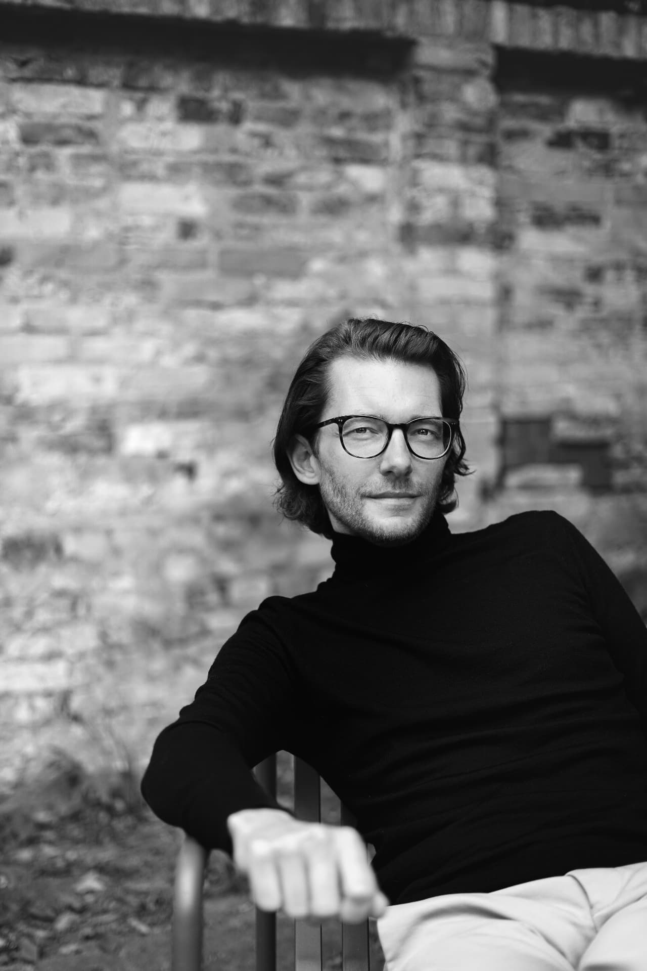 Black-and-white portrait of a man in glasses and a black turtleneck beside a brick wall