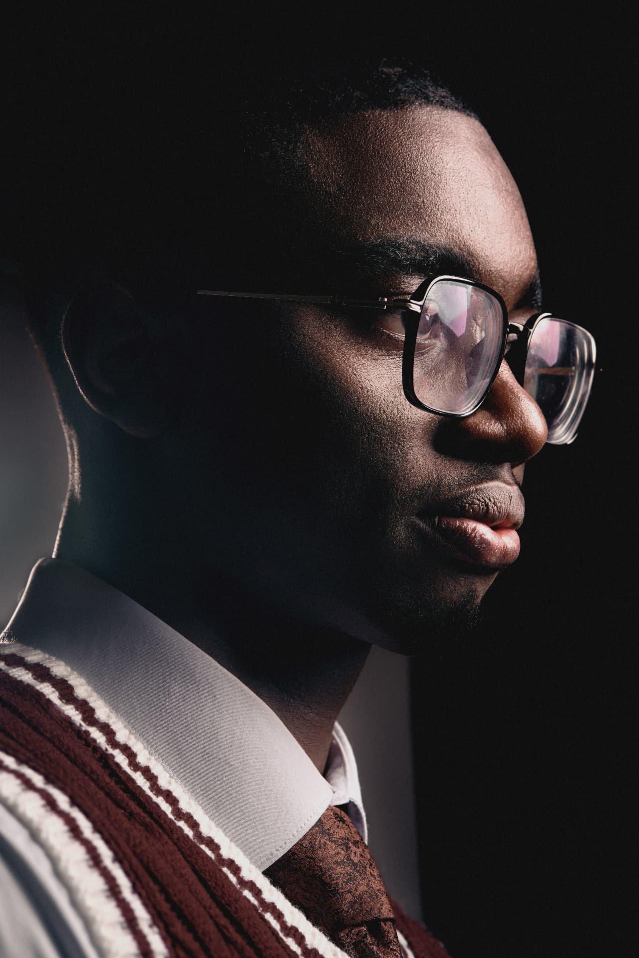 Profile portrait of a man in glasses, white shirt and patterned tie against a dark background